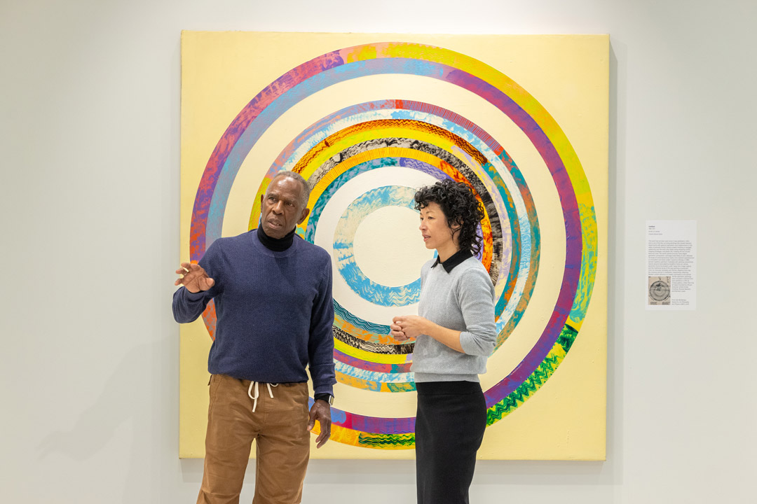 a man and a woman stand in an art gallery, with the man gesturing as explaining a piece of art beyond the field of view.