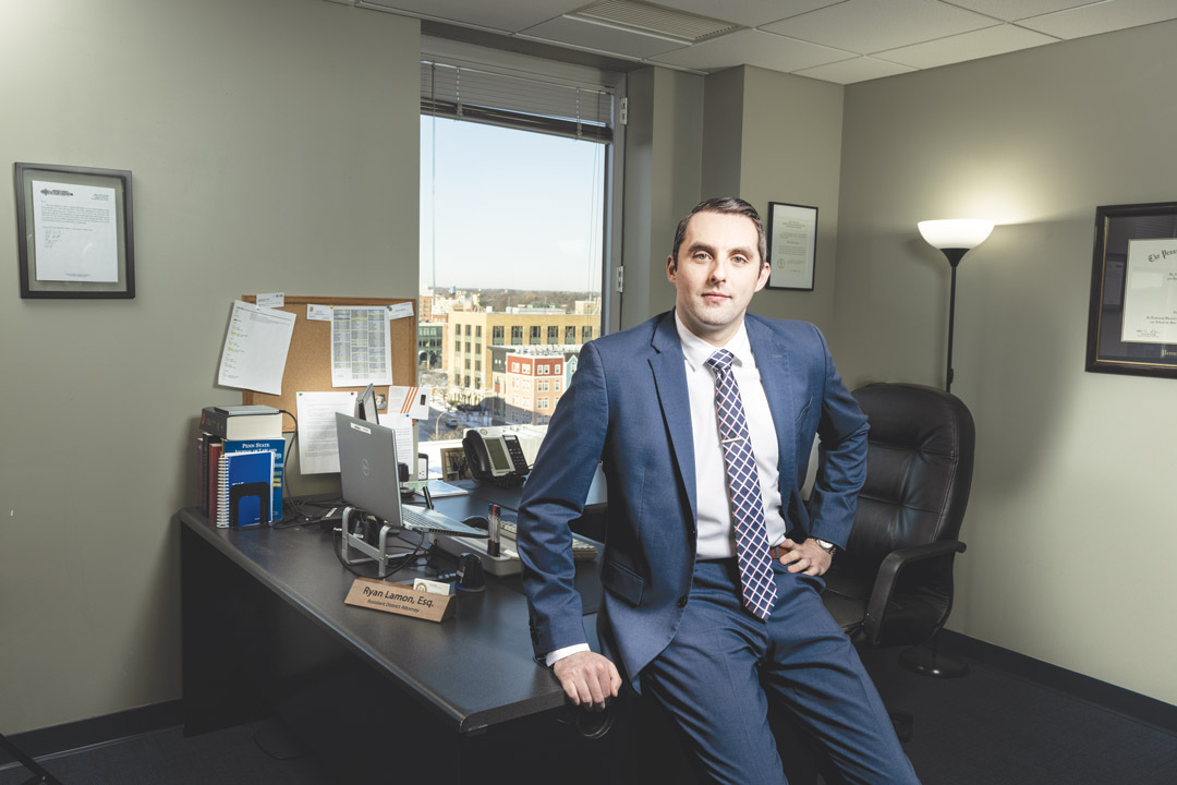 a man in a blue shirt leans on a desk in an office with a window looking out in to a city view.
