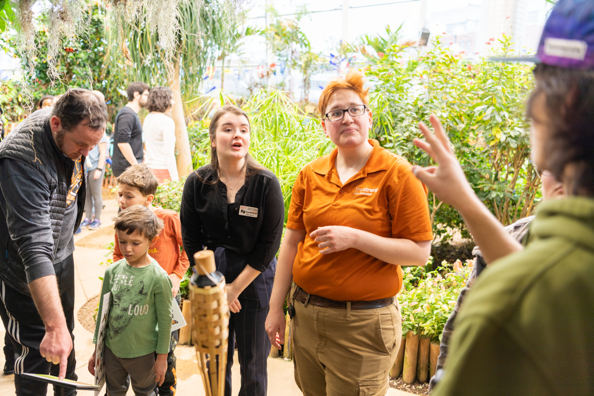 A student interpreter in a black shirt voices for a deaf student in a baseball cap and green hoodie as they ask questions of a butterfly expert in an orange shirt at The Strong Museum's Butterfly Garden. There are families with small children and lush greenery in the background. 