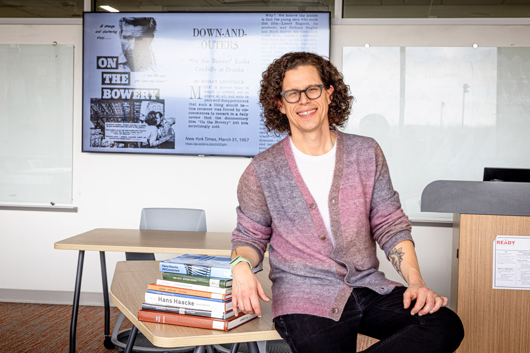 a man with a pink cardigan and shoulder length curly hair wears glasses and leans on a stack of books placed on a desk in a classroom.