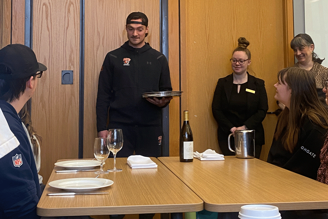 three students stand in front of a table where two students are seated with a table setting in front of them.