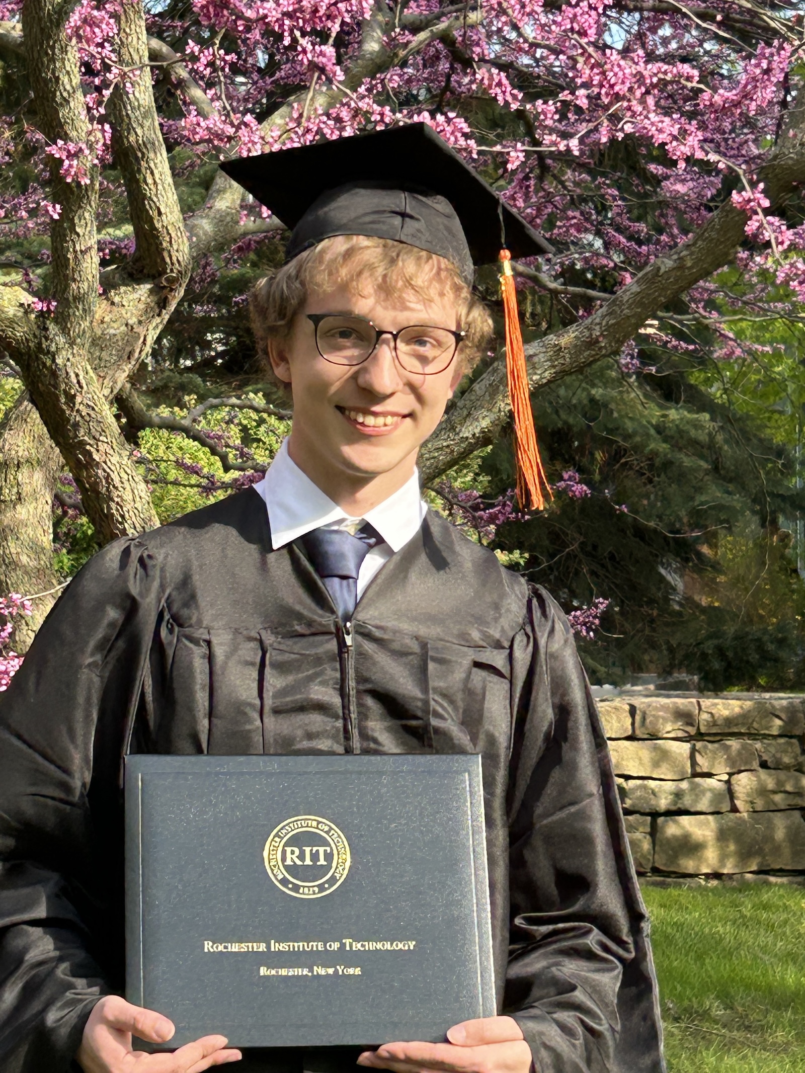 Student posing in front of flowering tree holding diploma, dressed in cap and gown