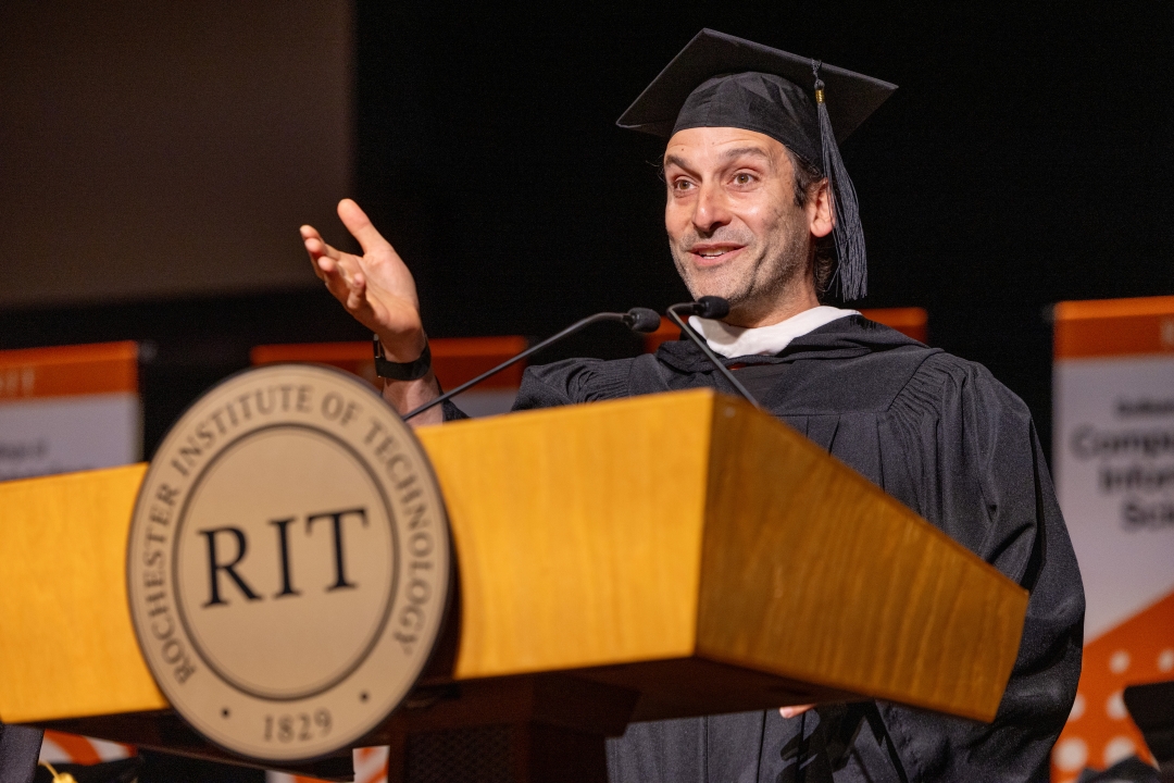 Man in black graduation cap and gown stands at podium with the R.I.T. logo.