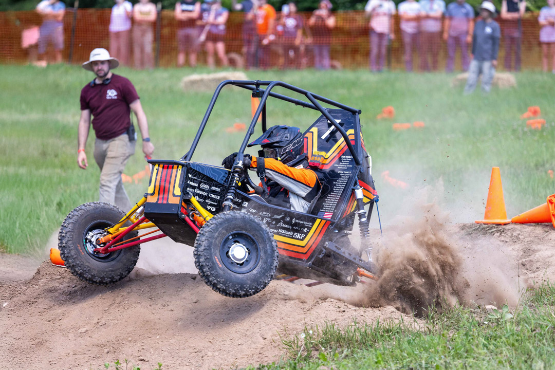 Baja SAE vehicle navigates dirt course, kicking up dust during competition.