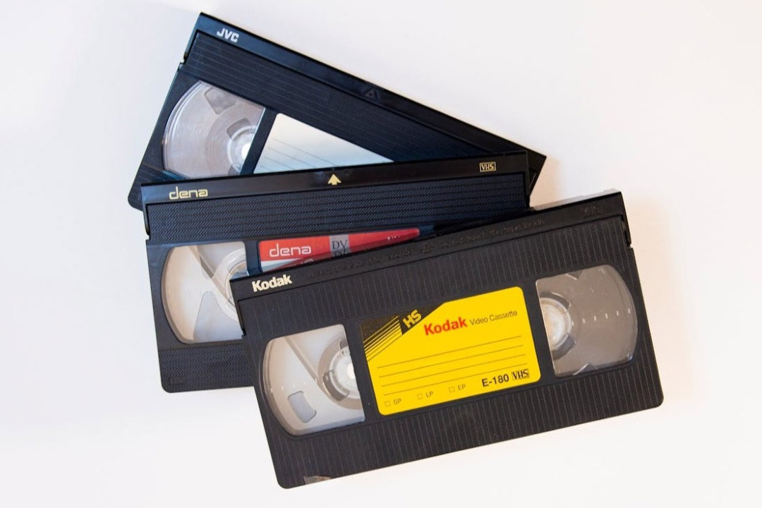 a pile of VHS tapes branded as Kodak, Dena, and JVC sit on a white tabletop