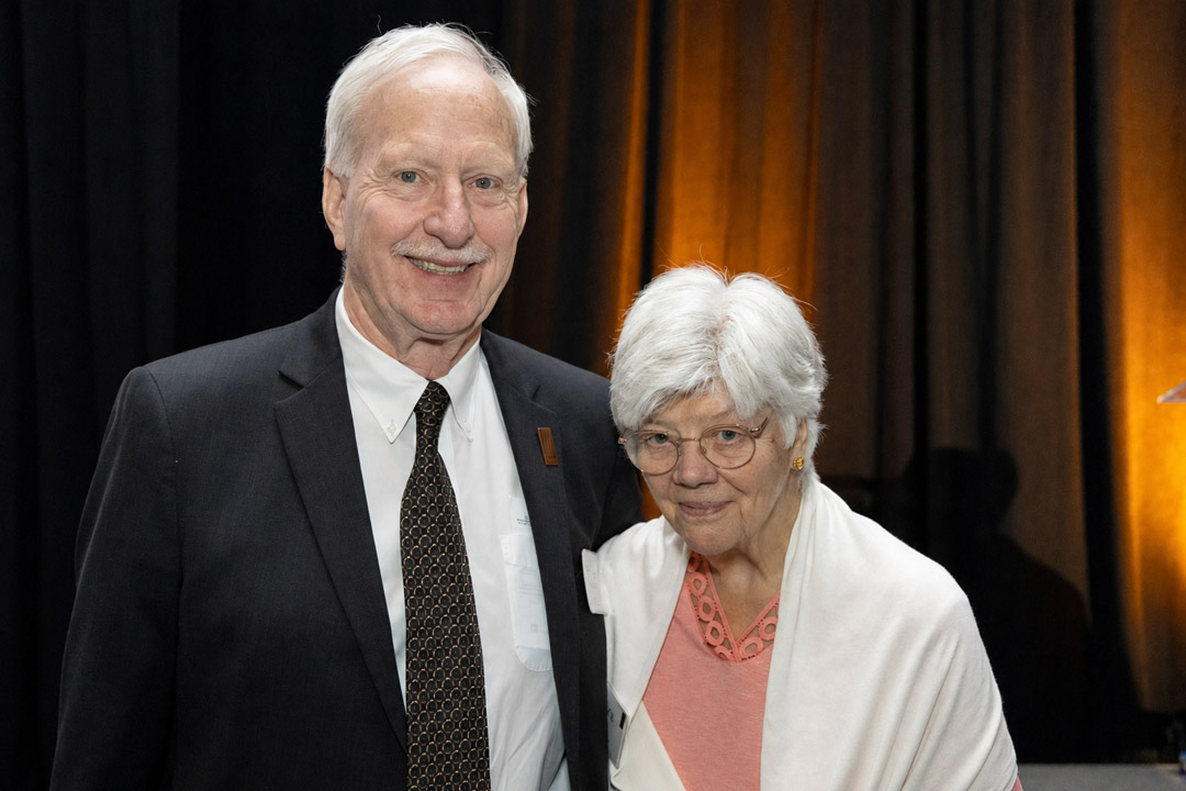 An older couple poses together at a formal event. The man wears a dark suit and patterned tie, while the woman wears a pink top with a white shawl. They stand in front of black drapes illuminated with warm amber lighting and smile toward the camera.