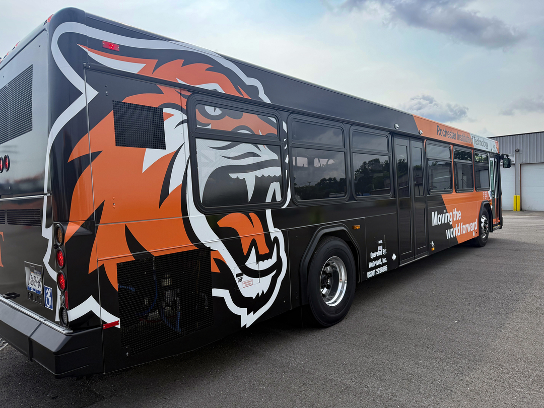 An image of the new RIT Bus. Looking at the side with the rear closest - it is black with an orange vertical stripe near the front and a tiger logo near the rear.