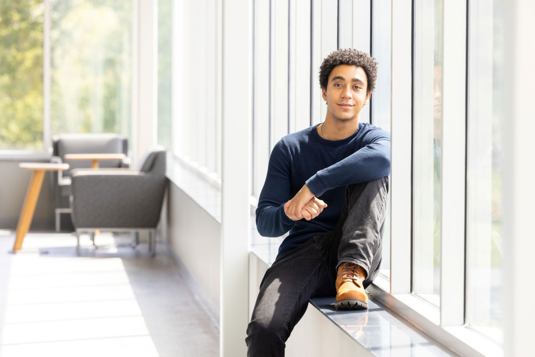 a college age male sits against a window in a brightly lit atrium with casual seating in the background.