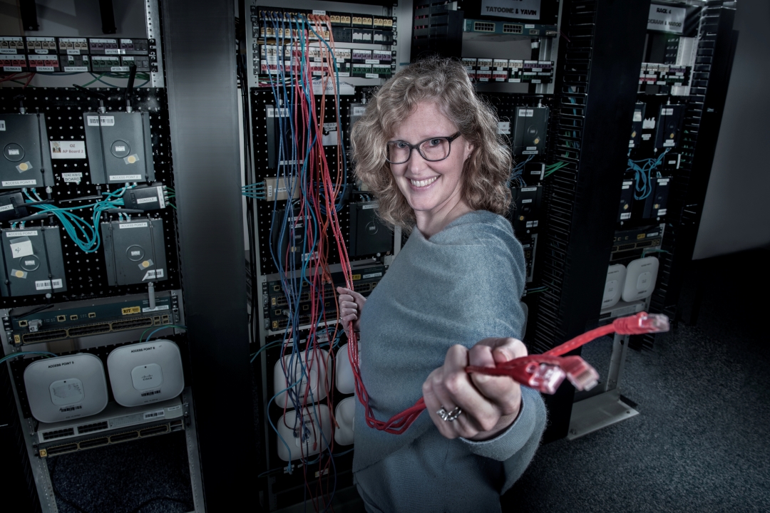Woman with glasses stands next to computer racks and holds red networking cables. 