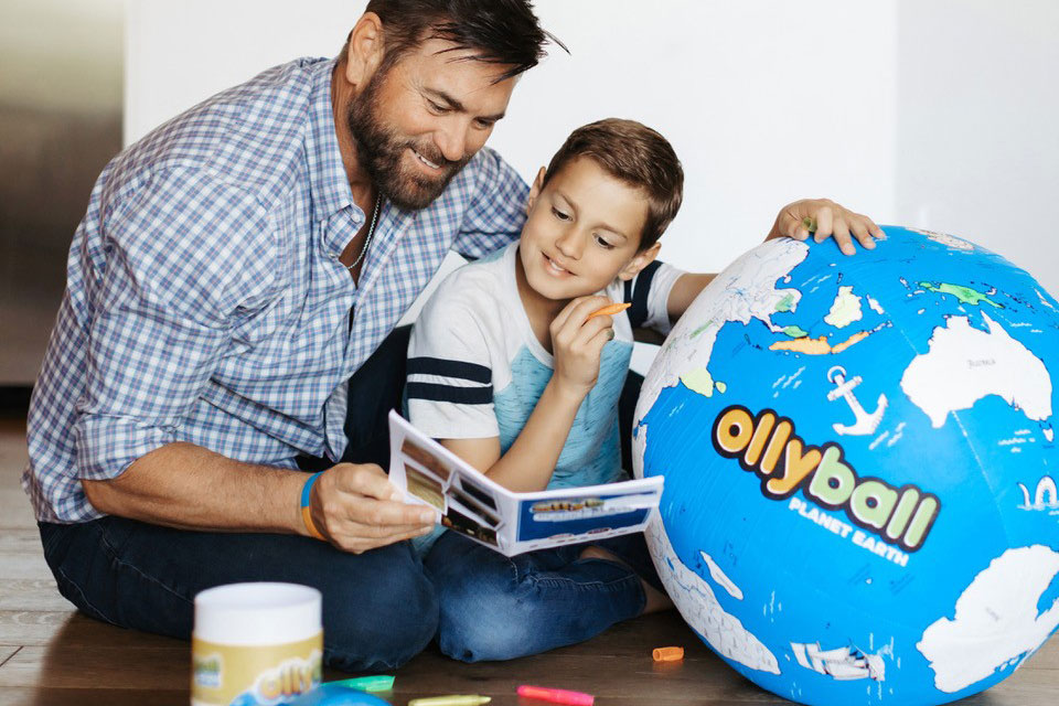 a man and a young boy sit on the floor next to a large ball designed to look like a world map that reads Ollyball on the side.