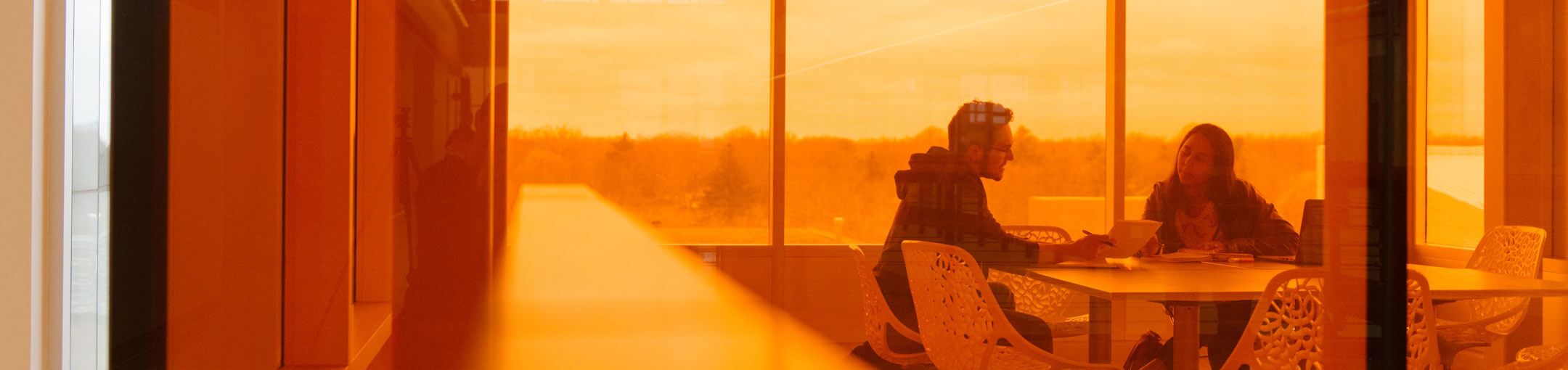 A man and woman sitting in a conference room looking over a paper with orange lighting.