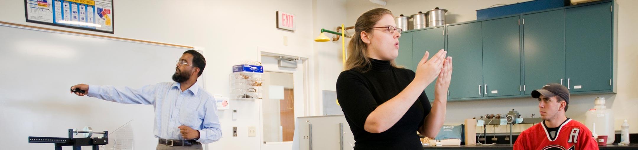 A male and female professor talk and point to a whiteboard in a lab while a male student observes.