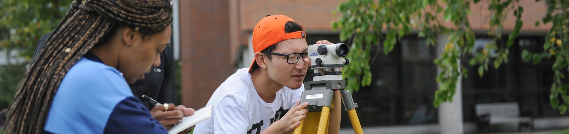 A student looks into camera while another student takes notes outside brick building.