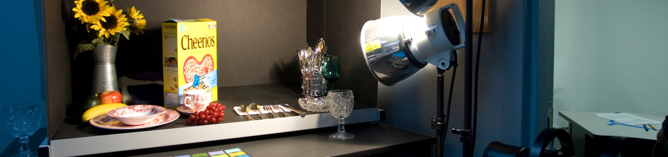 Desk with spotlight on plate, sunflowers in vase, utensils, glasses, and Cheerios cereal.