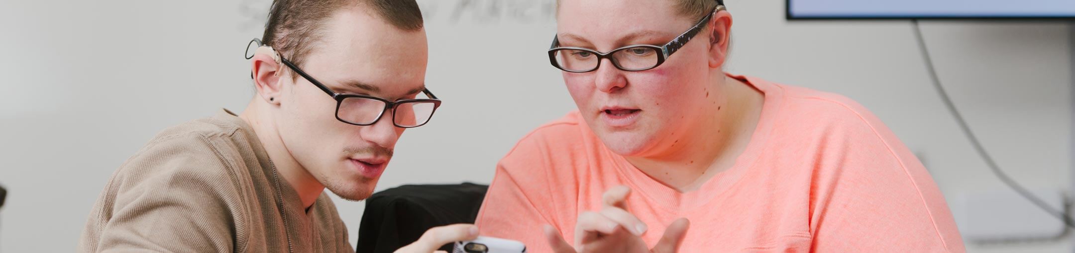 Two students, both wearing glasses, look at a smartphone.