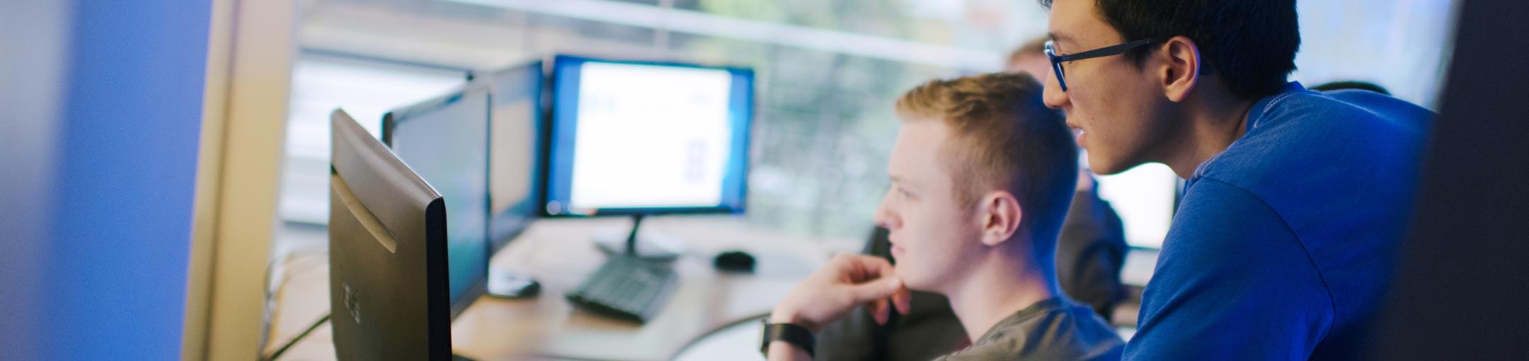 A sitting male students and another standing male student look at computer monitors.