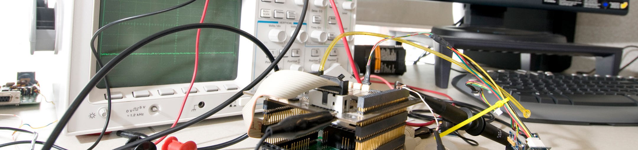 Circuit board, cords, and other electrical equipment with monitor and keyboard in background.