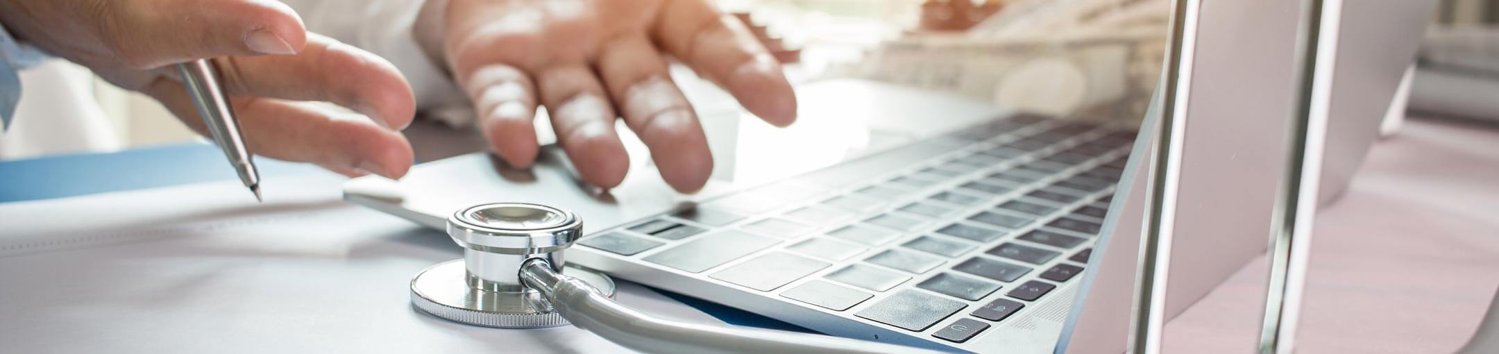 Hands holding pen work on laptop and stethoscope.