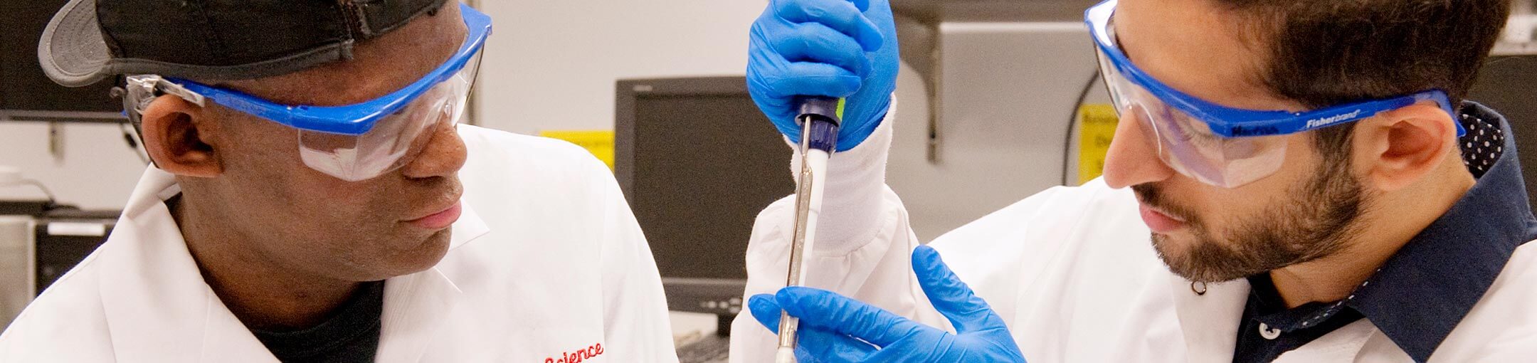 Two students wearing lab coats and protective goggles look at liquid in a pipette.