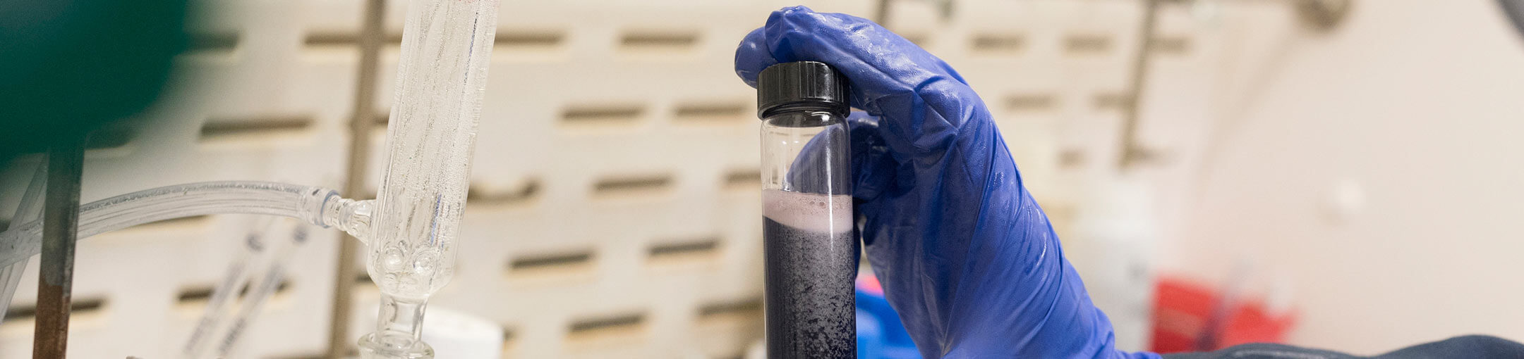 Blue gloved hands hold a capped tube of fizzy, dark liquid.