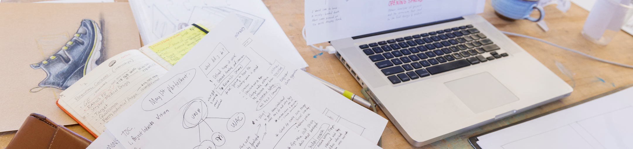 Close up of a laptop, notebook, and several pieces of paper with writing and diagrams.