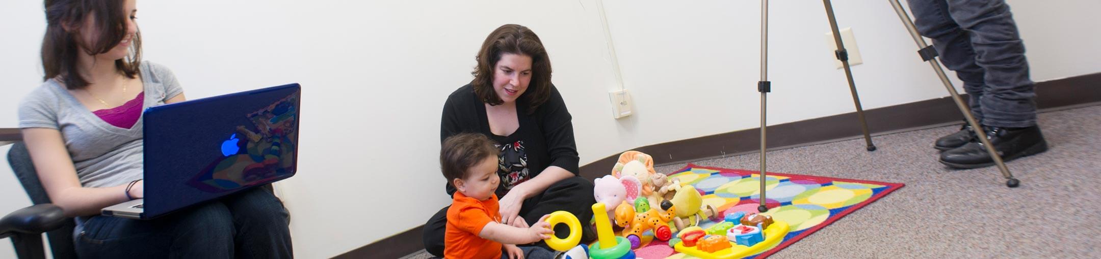 A person sits on a colorful play mat with a toddler while a student with a laptop watches.