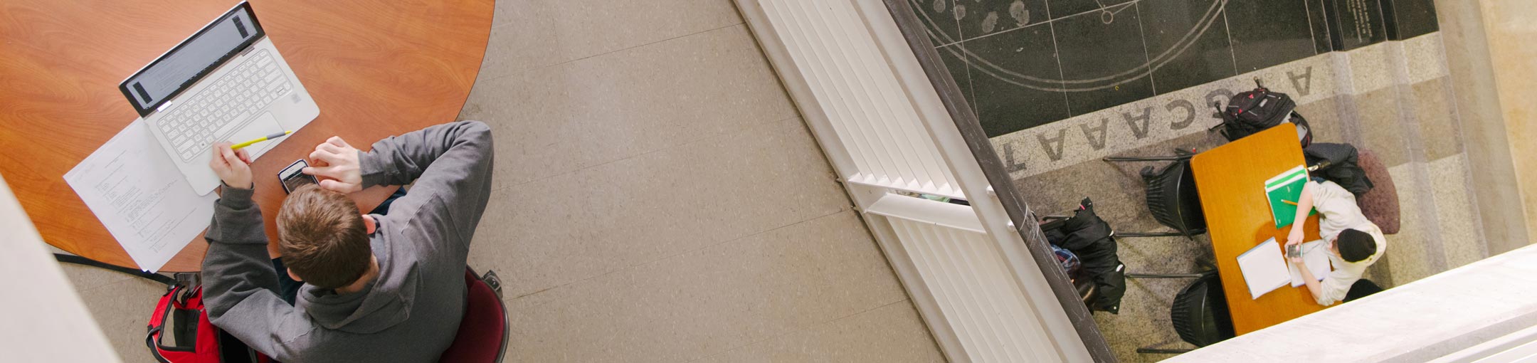 top-down view of a student working at a laptop on a balcony, with another student working at a table down a floor below.