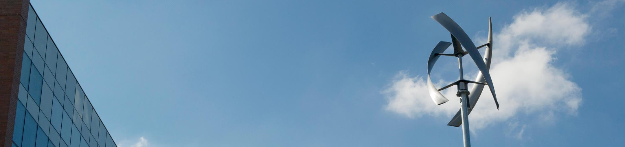 A vertical wind turbine sits in front of a building and a blue sky.