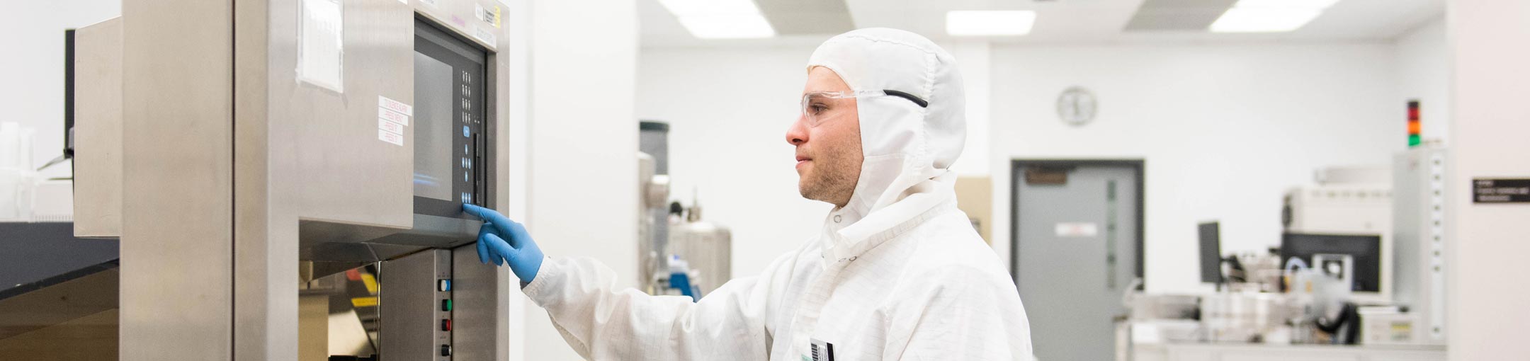 A student wearing a cleanroom suit presses a button on lab equipment