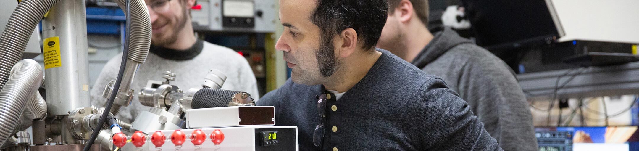 A student works in a lab with science equipment while two other students watch.