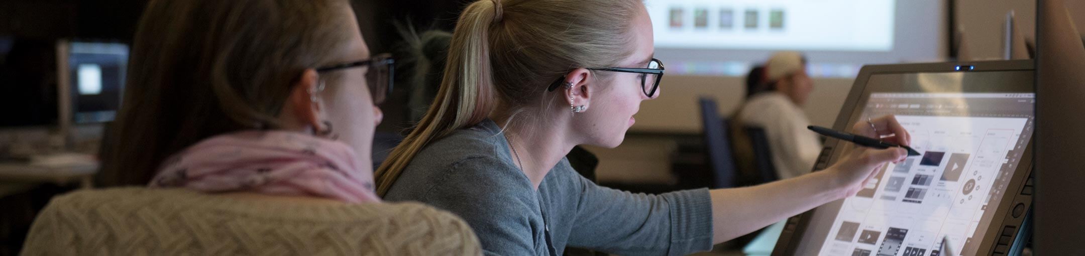 A student works on a large touchscreen monitor while another student watches.