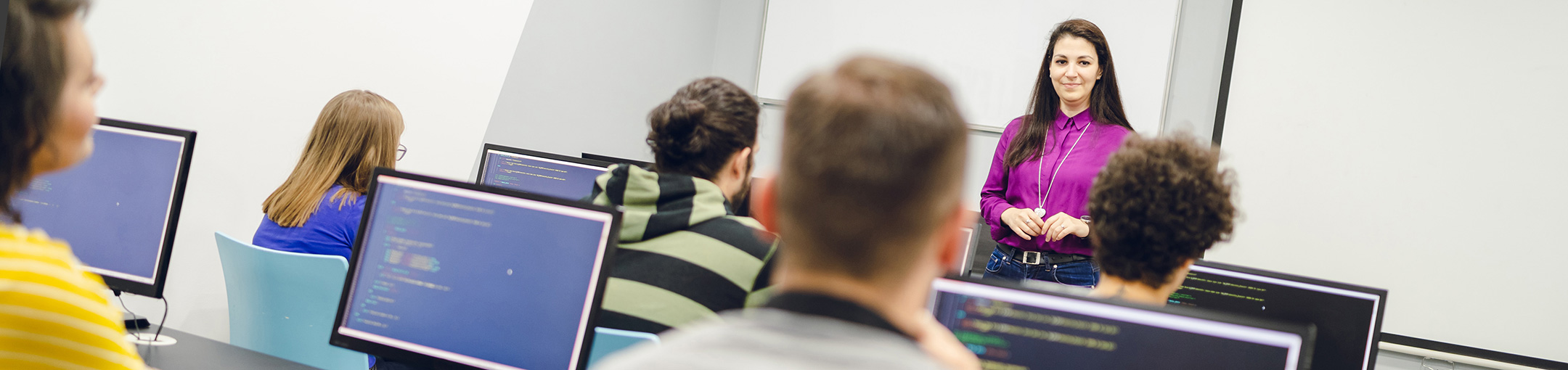 Female instructor teaching a class of students sitting at computers with code on screen