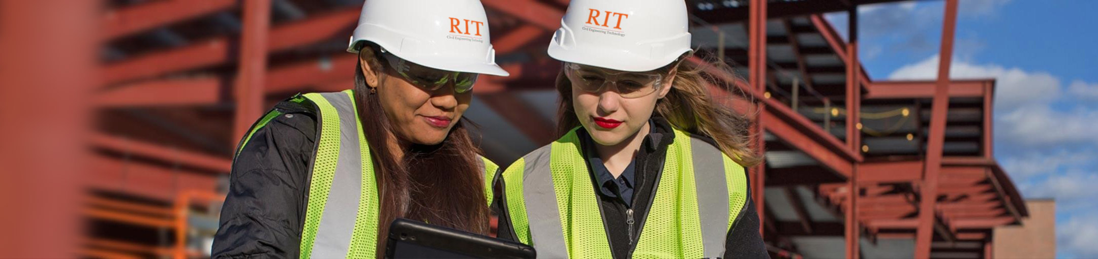 Two women wearing hard hats that say RIT look down at tablet computers. They are standing in from of a construction site.