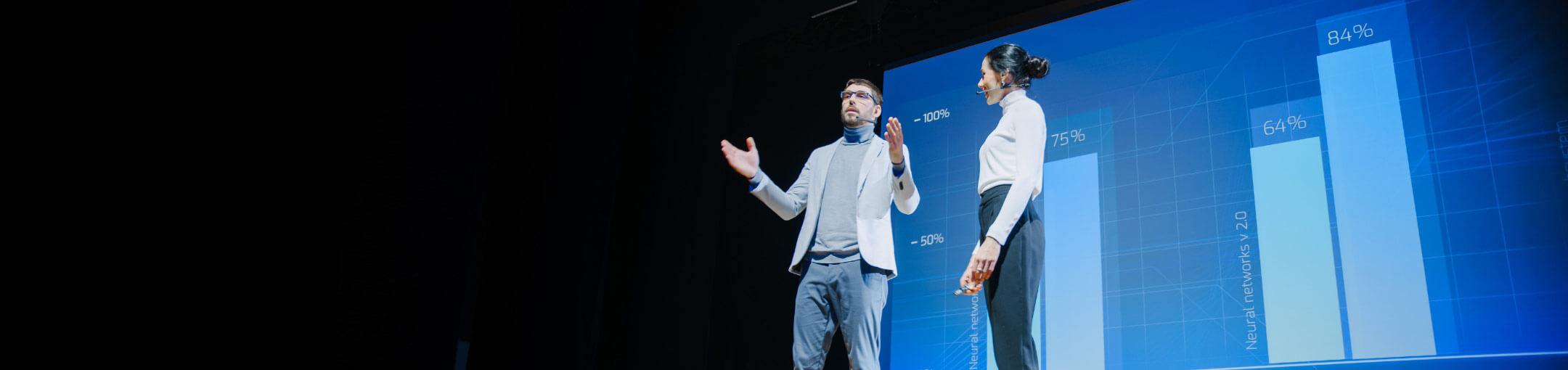 A man and woman stand on stage with charts projected behind them.