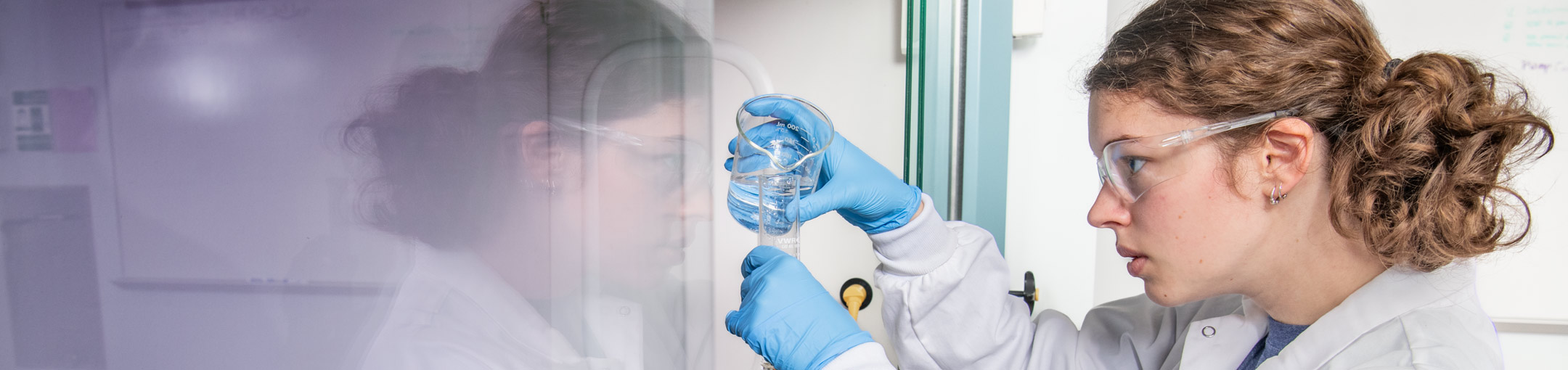 A student in a lab pours clear liquid into a beaker.