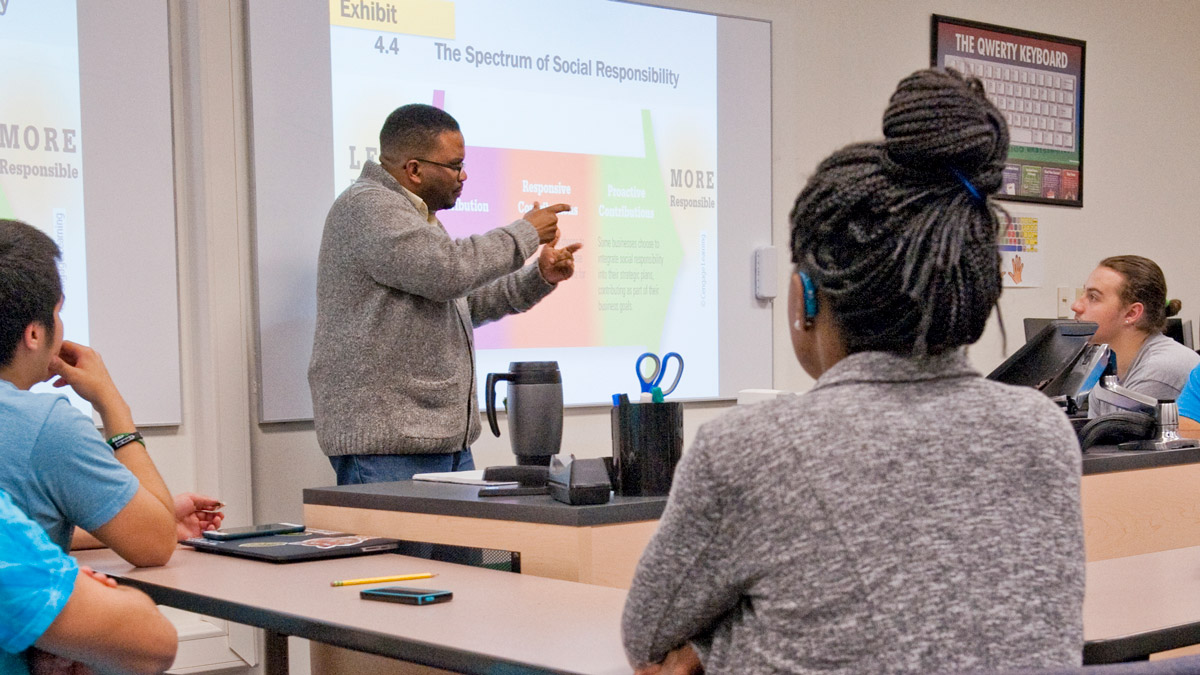 Professor standing in front of projector talks to a group of students in a classroom.