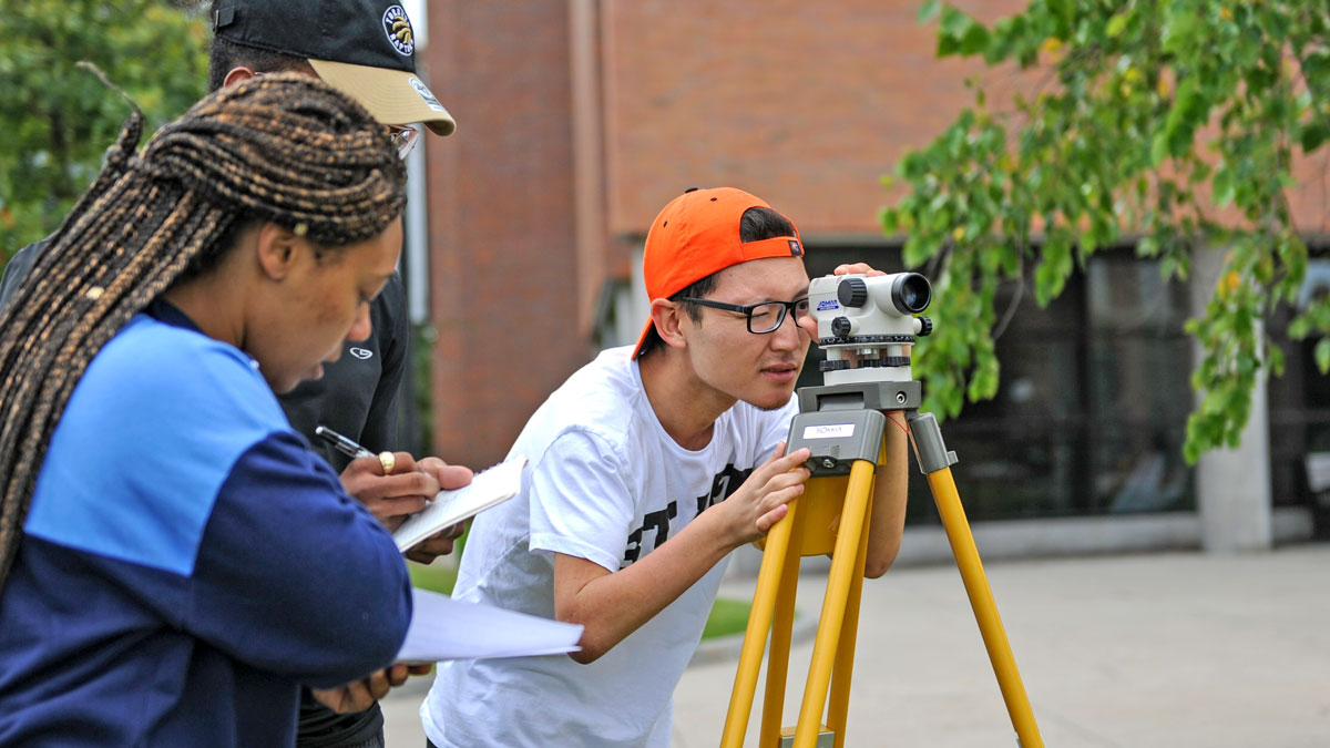 A student looks into surveying scope while other students take notes outside brick building.