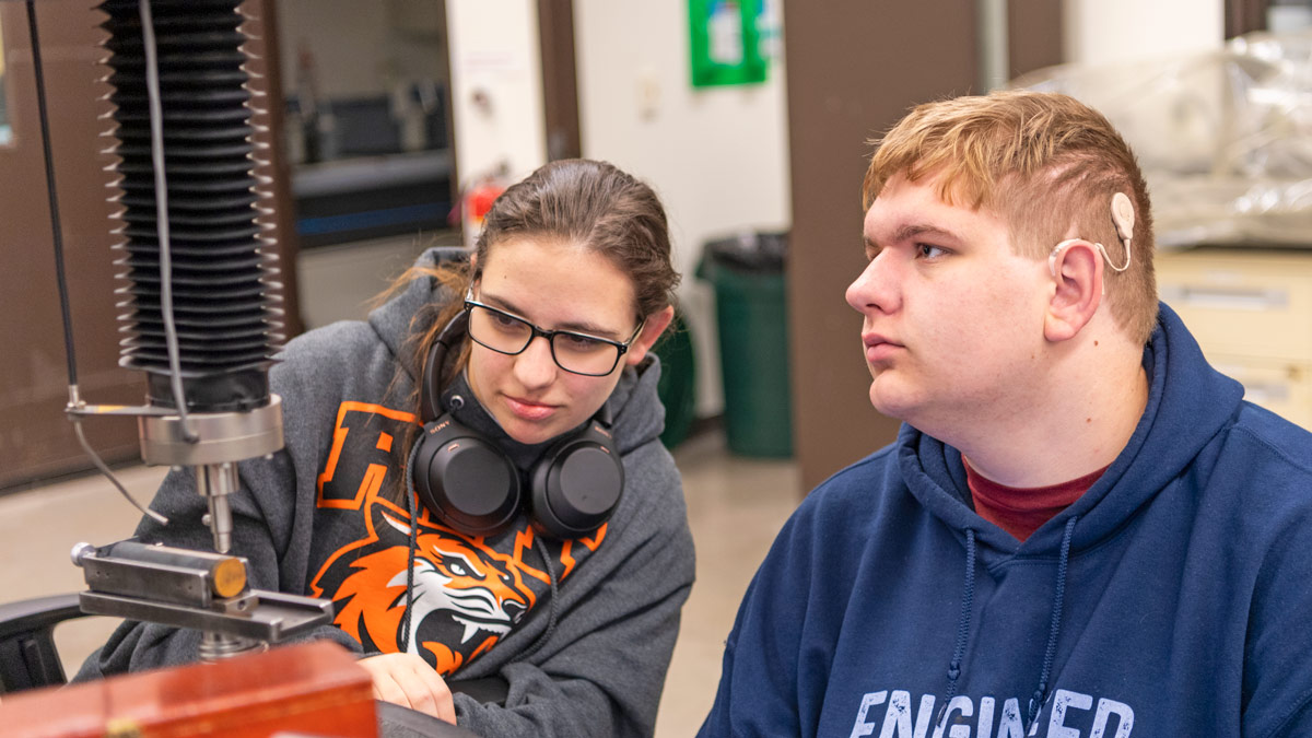 Two students sit in front of a machining device.