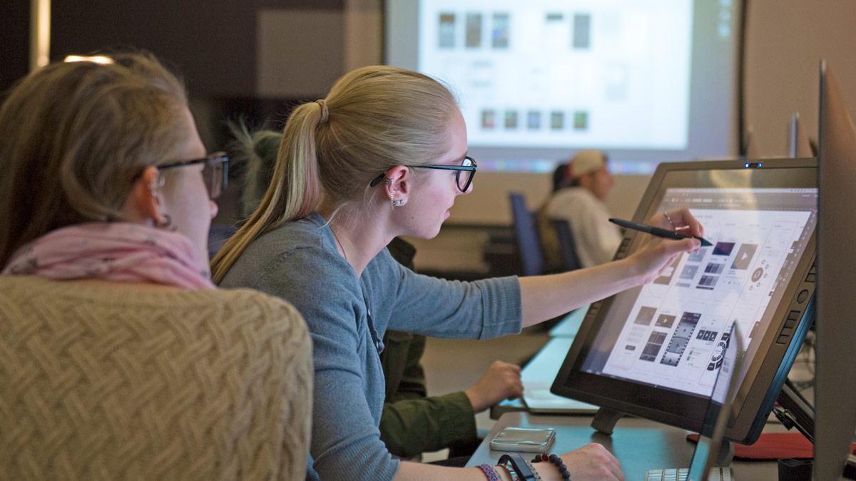 Two students with long hair and glasses work on a touchscreen computer monitor.