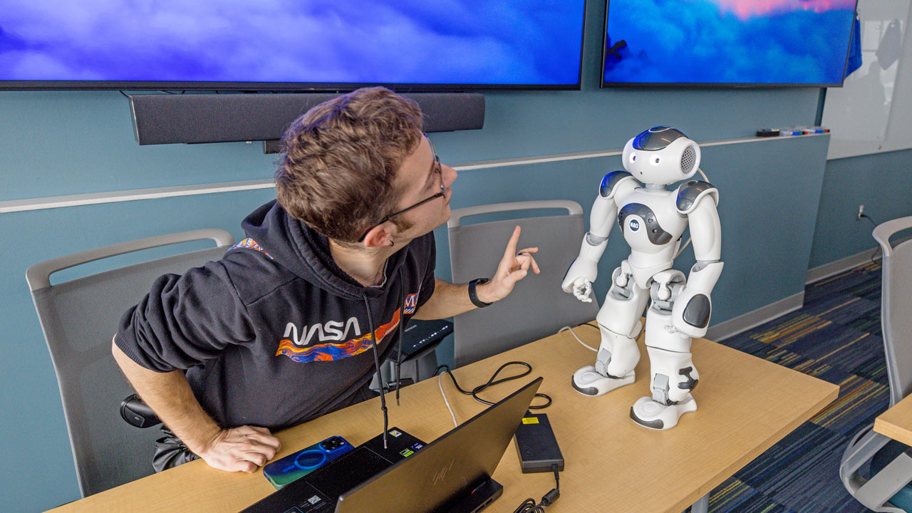 A student sits at a desk with a white 1-foot tall robot and a laptop computer.