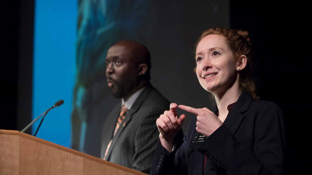 An ASL interpreter signs in the foreground as the presenter speaks at a podium in the background.
