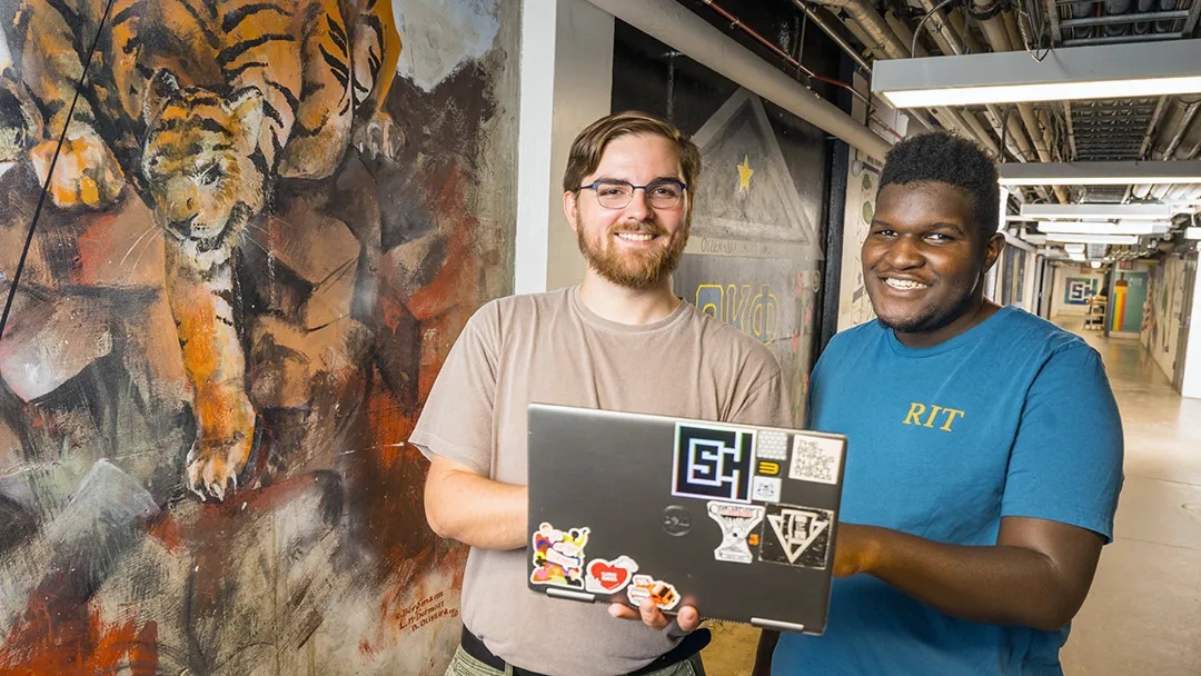 Two students with a laptop stand next to a mural.