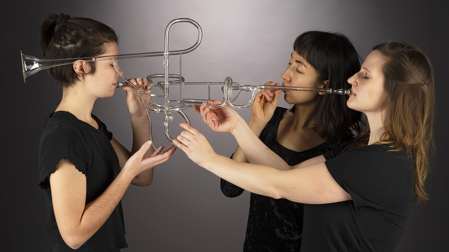 Three people play a glass trumpet that makes noise similar to a brass trumpet. The piece was designed to be an interactive art and music experience.