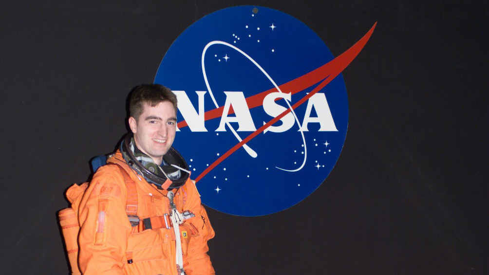 Paul Reichert stands in a space suit in front of a NASA wall decal.