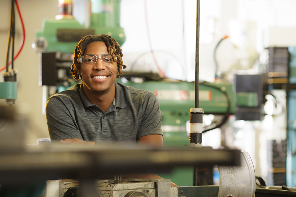 A RIT student engineer, wearing safety glasses and a gray collared shirt, looking at the camera and sitting in the Machine Shop.