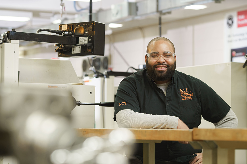 An mechanical engineering student wearing a black, button up shirt, sitting at a table in the machine shop.