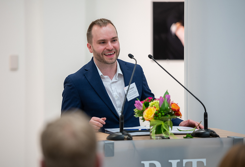 Matt Hartensveld speaking at a podium, wearing a navy blazer and a white shirt.