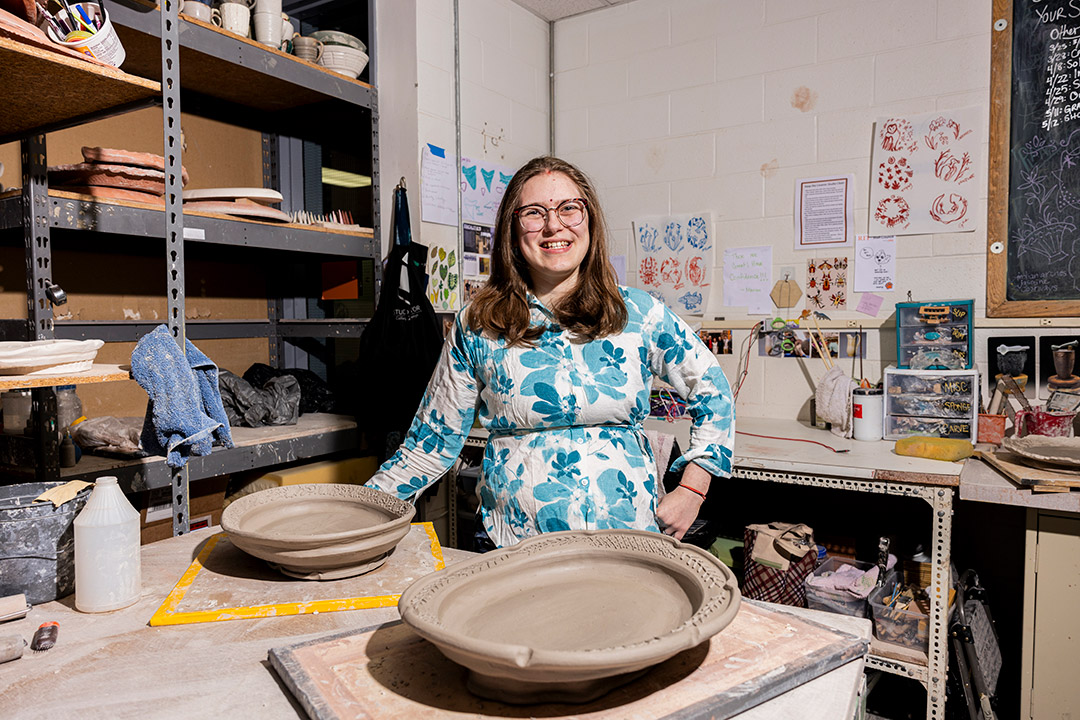 Emma Herz Thakur, who double majored in ceramics and illustration, stands before numerous ceramics art pieces in a studio. 