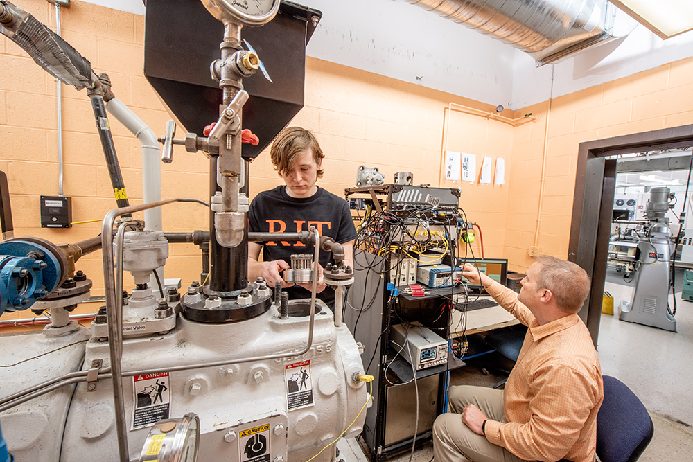 Jacob Chesnes working on a compressor while Dr. Jason Kolodziej works with wires in the background.