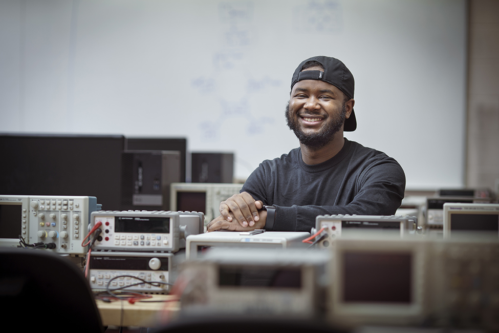 A college student looking at the camera and sitting in an electrical engineering lab in front of a whiteboard. 